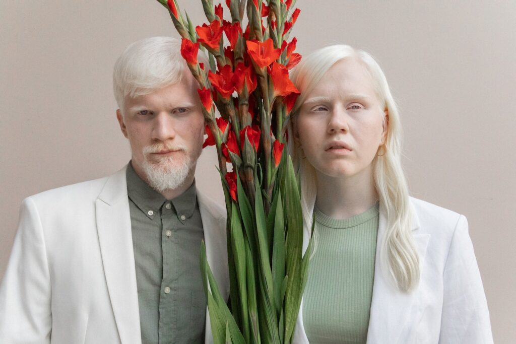 Charismatic couple in trendy outfits standing with bunch of red flowers with lush leaves and looking at camera while standing against beige background